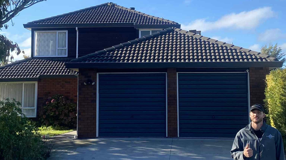 Person standing in front of a house with dark roof and garage doors.